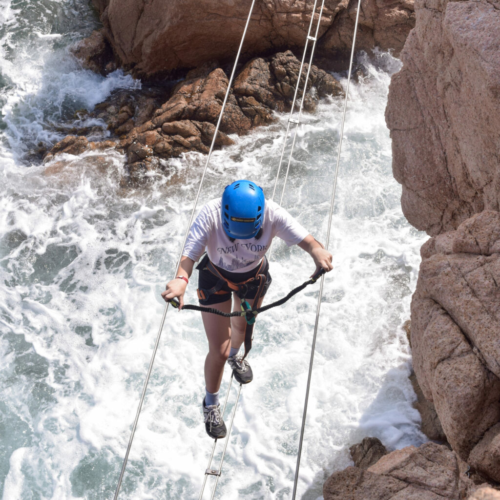 Dziewczyna przechodząca przez most linowy zawieszony nad falami. To Via Ferrata Cala Moli